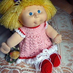 Könnte beinhalten: Ein rosa Häkelkleid mit weißen Verzierungen und roten Akzenten, getragen von einer Puppe mit blonden Garnhaaren und blauen Augen.