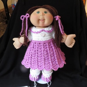 May include: A handmade doll with brown pigtails tied with purple ribbons. The doll is dressed in a white and purple crochet dress with a heart, white socks, and purple shoes. The doll has green eyes and rosy cheeks, a classic toy.