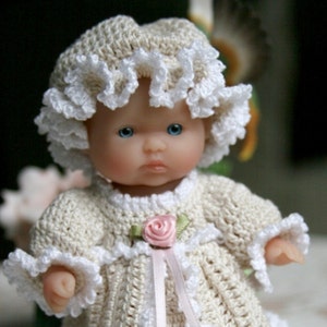 May include: A crocheted doll wearing a white crocheted bonnet and a cream crocheted dress with a pink flower accent.