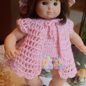 May include: A doll wearing a pink crocheted dress and matching cardigan with a colorful crocheted hat. The doll is wearing pink crocheted shoes and is standing on a wooden base.