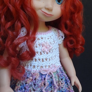 Może przedstawiać: Lalka z rudymi kręconymi włosami i niebieskimi oczami, ubrana w ręcznie robiony szydełkowy strój. Biały top ma różowy kwiat, a spódnica jest wielokolorowa. Lalka kolekcjonerska.
