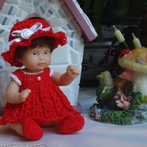 Op de afbeelding: Een rode gehaakte jurk met witte versiering en een bijpassende rode gehaakte hoed met een witte bloemenaccent. De pop draagt ​​de jurk en de hoed en zit op een wit oppervlak.