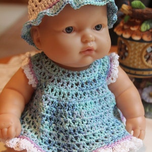Puede incluir: Una muñeca de crochet que lleva un sombrero de crochet blanco con ribete azul claro y morado. La muñeca lleva un vestido de crochet azul claro y morado con ribete de crochet blanco.