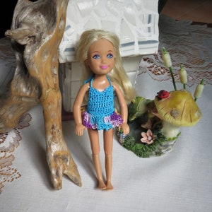 Puede incluir: Una muñeca rubia con un traje de baño de ganchillo azul con ribetes morados y rosas. La muñeca está de pie sobre una superficie blanca con una rama de madera y una figura de hongo en el fondo.