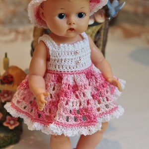 May include: A porcelain doll wearing a white and pink crocheted dress and hat. The doll has blue eyes and a pink and white crocheted hat.