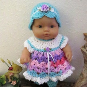 May include: A crocheted doll outfit in shades of pink, purple, blue, and white. The outfit includes a dress with a white crocheted collar and a matching hat with a flower embellishment.