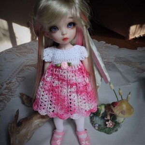Könnte beinhalten: Eine kleine Puppe mit blonden Haaren und blauen Augen, die ein rosa-weißes Häkelkleid trägt. Die Puppe steht auf einer Holzoberfläche mit einer kleinen Pilzfigur im Hintergrund.