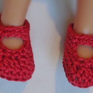 May include: A pair of handmade, bright red crochet baby shoes. The shoes have a Mary Jane style with a strap and a round opening on the top. The crochet stitch creates a textured appearance. The shoes are designed for a baby.