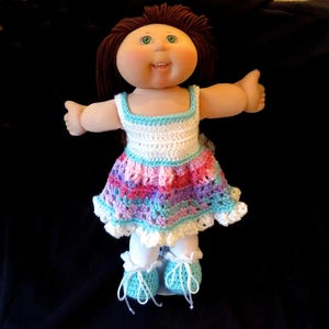 May include: A vintage doll with brown hair and green eyes, wearing a handmade crochet dress in white, pink, purple, and blue. The doll also has matching blue shoes with white laces. The doll is standing with its arms open.