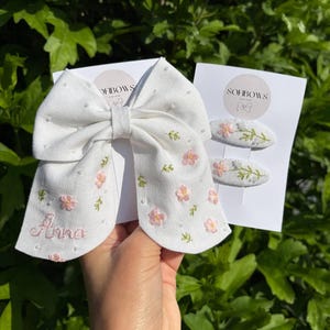 Può includere: Un fiocco per capelli in tessuto bianco e due fermagli ovali abbinati, tutti decorati con fiori rosa ricamati, steli verdi e piccole perline bianche. Il fiocco è personalizzato con il nome Anna ricamato in filo rosa su una delle code.