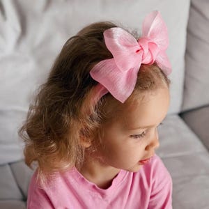 Puede incluir: Una diadema rosa con un lazo grande y transparente. El lazo está anudado en el centro y se coloca sobre la cabeza. La diadema y el lazo son de color rosa suave. La persona de la imagen lleva una camiseta rosa.