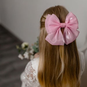 Puede incluir: Un gran lazo rosa claro como accesorio para el cabello. El lazo está hecho de una tela transparente y brillante, y está sujeto a la parte posterior de la cabeza. El cabello es largo y rubio. El lazo es el punto focal de la imagen.