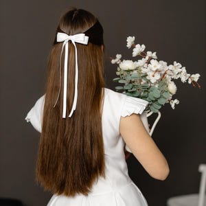 Può includere: Una giovane persona con lunghi capelli castani indossa un abito bianco con un fiocco di raso bianco nei capelli. Sta tenendo un mazzo di fiori bianchi.
