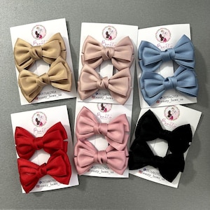 Pode incluir: Seis pares de laços de cabelo de cetim em várias cores, incluindo bege, rosa, azul, vermelho e preto. Cada par é exibido em um cartão branco com o texto "beauty bows ua" e um número de telefone.
