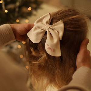 May include: A beige fabric bow with the name "Conner" embroidered in pink on the back. The bow is worn in a girl's hair.
