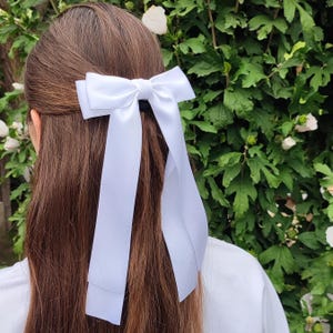 Könnte beinhalten: Eine weiße Haarschleife aus Satin mit langen Bändern ist im Haar befestigt. Die Schleife ist hinten am Kopf zentriert, wobei die Bänder über das braune Haar fallen. Der Hintergrund zeigt grünes Laub und weiße Blüten.