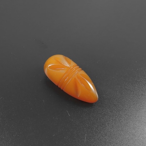 May include: A teardrop-shaped, amber-colored pendant with carved floral designs. The pendant has a smooth, polished surface and is set against a dark gray background. The carved details include lines and petal-like shapes, adding texture and visual interest.