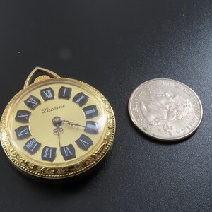 May include: A gold-toned Lucerne pocket watch with a cream-colored face and black Roman numerals. The watch is next to a U.S. quarter coin for size comparison. The watch has a decorative bezel and a loop for a chain.