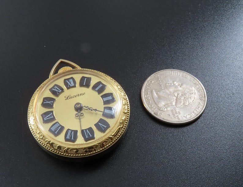 May include: A gold-toned Lucerne pocket watch with a cream-colored face and black Roman numerals. The watch is next to a U.S. quarter coin for size comparison. The watch has a decorative bezel and a loop for a chain.