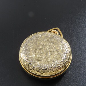 May include: A gold-toned pocket watch with an ornate, engraved floral design on the cover. The watch has a circular shape and a small loop at the top. A silver quarter coin is next to the watch.