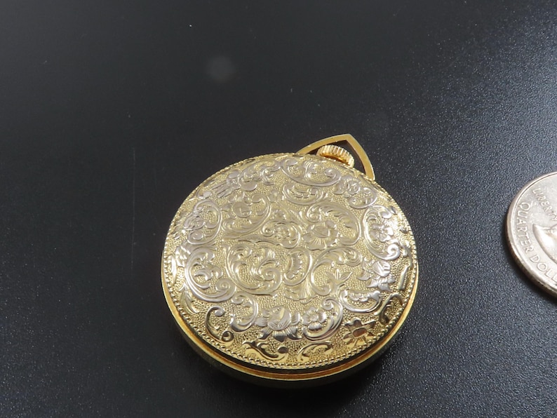 May include: A gold-toned pocket watch with an ornate, engraved floral design on the cover. The watch has a circular shape and a small loop at the top. A silver quarter coin is next to the watch.