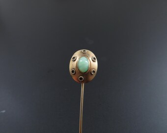 May include: A vintage hat pin with a gold-toned oval head. The head features a light green oval stone in the center, surrounded by small, dark accents. The pin is a classic accessory.