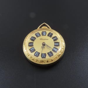 May include: A gold Lucerne pocket watch with a cream-colored face and black Roman numerals. The watch has a decorative gold bezel and a small loop at the top for a chain. The watch hands are black.