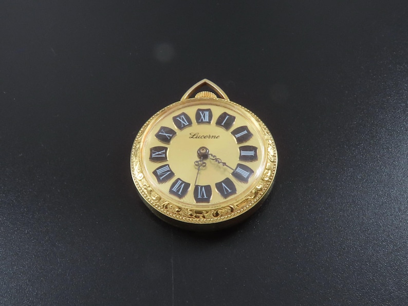 May include: A gold Lucerne pocket watch with a cream-colored face and black Roman numerals. The watch has a decorative gold bezel and a small loop at the top for a chain. The watch hands are black.