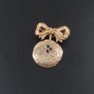 May include: A gold-toned metal brooch featuring a bow at the top connected to a circular pendant. The pendant has an embossed swirling pattern and is accented with four small, colorful round stones in green, blue, purple, and red at the center.
