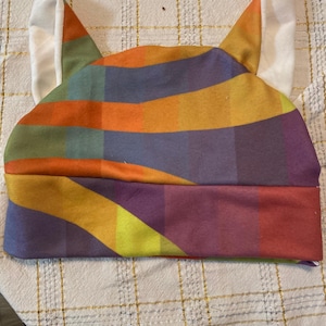 May include: A colorful, striped hat with cat ears. The hat features a vibrant pattern of orange, yellow, purple, and blue stripes. The ears are white and stand upright. The hat is made of soft fabric.