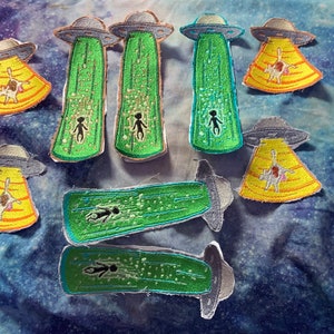 May include: A collection of 14 embroidered patches featuring a green alien spaceship abducting a cow. The patches are on a blue and purple fabric background.