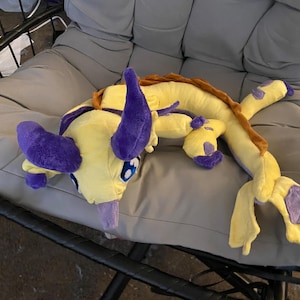 May include: A plush toy dragon in yellow, purple, and brown. The dragon has large purple horns, blue eyes, and a long tail. It is lying on a gray cushion. The toy is soft and cuddly, perfect for children.