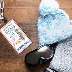 Puede incluir: Un pase de esquí rectangular con borde rosa, código de barras y el texto ALLIE'S APRES SKI, SKI LIFT PASS, ALL DAY ACCESS y VALID THRU 4/5/2025. El pase está unido a un cordón blanco junto a un gorro azul claro con pompón, gafas de esquí negras y un suéter gris.