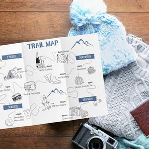 Op de afbeelding: Een drievoudig gevouwen papieren routekaart met blauwe en zwarte illustraties en tekst die een weekendplanning weergeeft. De kaart wordt vergezeld door een lichtblauwe gebreide muts met pompon, een grijze kabeltrui, een vintage camera en een bruin paspoort.