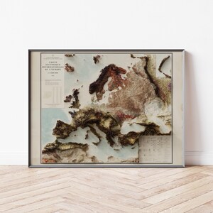 Puede incluir: Mapa en relieve 3D enmarcado de Europa, que muestra la topografía del continente en tonos marrones, verdes y beige. El mapa incluye texto en francés y un fondo oceánico azul claro. La obra de arte se exhibe en una pared blanca.