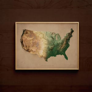 May include: Framed topographic map of the United States, showcasing the country's varied terrain in shades of brown and green. The map is set against a neutral background and framed in a gold-colored border, mounted on a dark wood surface.