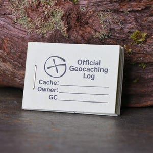 May include: A small, white, paper logbook for geocaching. The cover features a black compass symbol and the text "Official Geocaching Log". The logbook has lines for "Cache:" and "Owner:" and a space for a "GC" code.