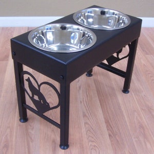 Pode incluir: Suporte de tigelas para comida e &aacute;gua para c&atilde;es em metal preto com duas tigelas de a&ccedil;o inoxid&aacute;vel. O suporte apresenta um recorte em forma de cora&ccedil;&atilde;o com uma silhueta de c&atilde;o.