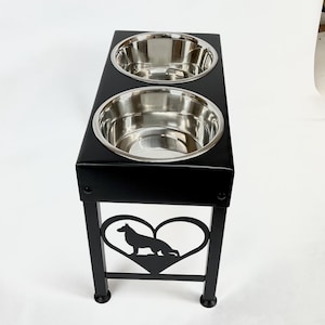 Pode incluir: Suporte para tigelas de c&atilde;o em metal preto com duas tigelas de a&ccedil;o inoxid&aacute;vel e um recorte em forma de cora&ccedil;&atilde;o com uma silhueta de pastor alem&atilde;o.