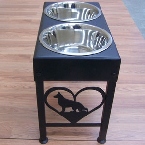 Pode incluir: Suporte para comida de c&atilde;o de metal preto com duas tigelas de a&ccedil;o inoxid&aacute;vel e um recorte em forma de cora&ccedil;&atilde;o com a silhueta de um pastor alem&atilde;o.