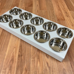 May include: White metal tray with ten stainless steel cups. The tray is rectangular and has a smooth surface. The cups are evenly spaced and have a shiny finish.