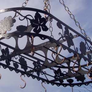 May include: Black metal pot rack with a grapevine design. The rack has multiple hooks for hanging pots and pans. The rack is suspended from the ceiling by a chain.
