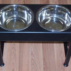 Pode incluir: Suporte de tigela dupla para c&atilde;es de metal preto com duas tigelas de a&ccedil;o inoxid&aacute;vel. O suporte tem quatro pernas e &eacute; elevado do ch&atilde;o.