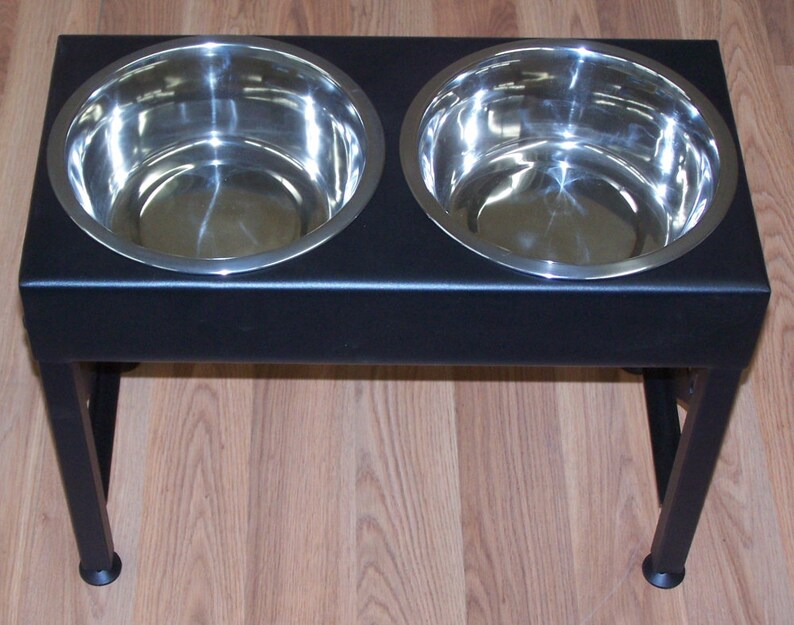 Pode incluir: Suporte de tigela dupla para c&atilde;es de metal preto com duas tigelas de a&ccedil;o inoxid&aacute;vel. O suporte tem quatro pernas e &eacute; elevado do ch&atilde;o.