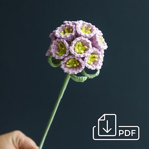 May include: A handmade crochet flower with a cluster of lavender-colored blooms, each with a bright green center. The flower has a green stem and leaves. The craft project is set against a dark background.