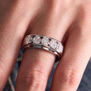 May include: A silver ring with six round, clear gemstones set in a channel. The ring is on a finger, with a denim background visible.