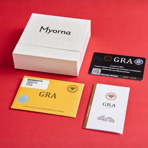 May include: A white textured box with the brand name "Myorna" in black lettering, accompanied by a black warranty card and two certificates on a red surface. One certificate is yellow with "GRA" printed on it.