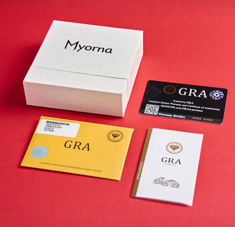 May include: A white textured box with the brand name "Myorna" in black lettering, accompanied by a black warranty card and two certificates on a red surface. One certificate is yellow with "GRA" printed on it.