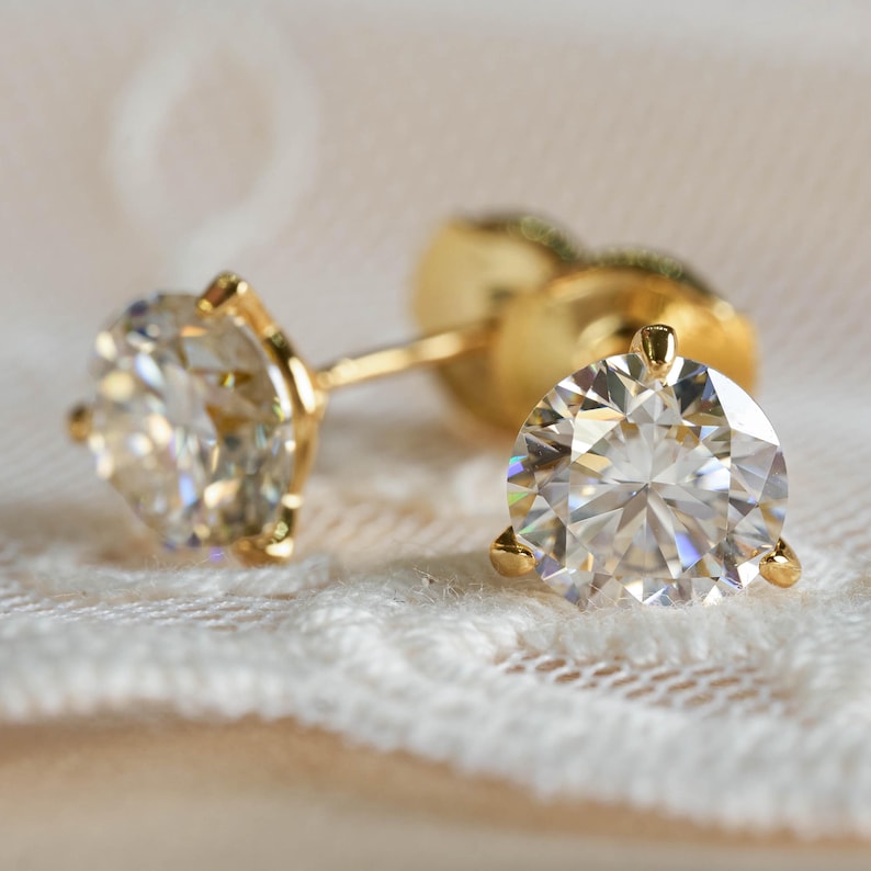 May include: Close-up of two gold stud earrings with large, round, clear gemstones. The earrings are set in a classic three-prong setting, showcasing the brilliance of the stones. The background is a soft, blurred fabric.
