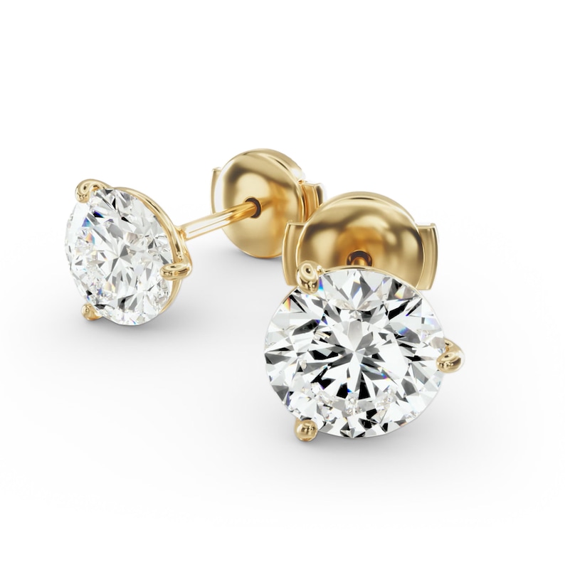 May include: A pair of gold stud earrings with large, round, clear gemstones. The earrings have a classic three-prong setting and a post and butterfly backing. The gold has a polished finish, and the gemstones sparkle brightly against the white background.